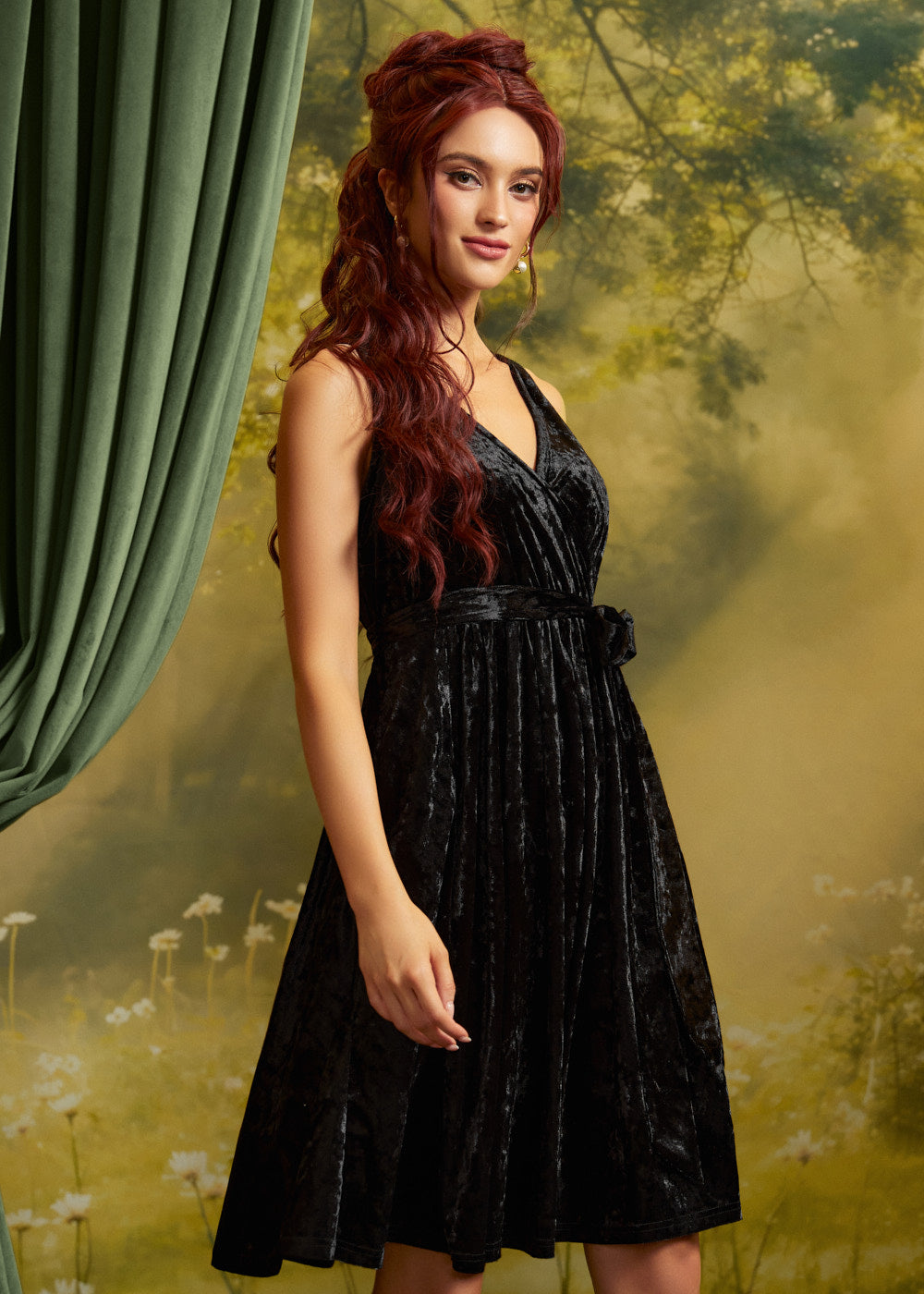 A woman with long wavy red hair models a sleeveless black velvet midi dress with a V-neckline and matching waist tie, posed against a moody, misty forest backdrop with a green curtain on the left.