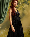 A woman with long wavy red hair models a sleeveless black velvet midi dress with a V-neckline and matching waist tie, posed against a moody, misty forest backdrop with a green curtain on the left.