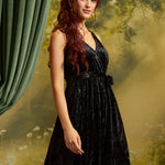 A woman with long wavy red hair models a sleeveless black velvet midi dress with a V-neckline and matching waist tie, posed against a moody, misty forest backdrop with a green curtain on the left.