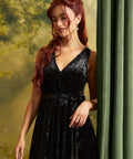 A woman with long wavy red hair models a sleeveless black velvet wrap dress with a tie waist, standing next to a green curtain against a soft yellow-green backdrop.