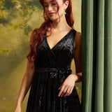 A woman with long wavy red hair models a sleeveless black velvet wrap dress with a tie waist, standing next to a green curtain against a soft yellow-green backdrop.