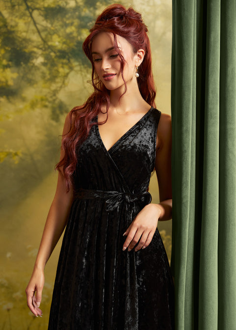 A woman with long wavy red hair models a sleeveless black velvet wrap dress with a tie waist, standing next to a green curtain against a soft yellow-green backdrop.