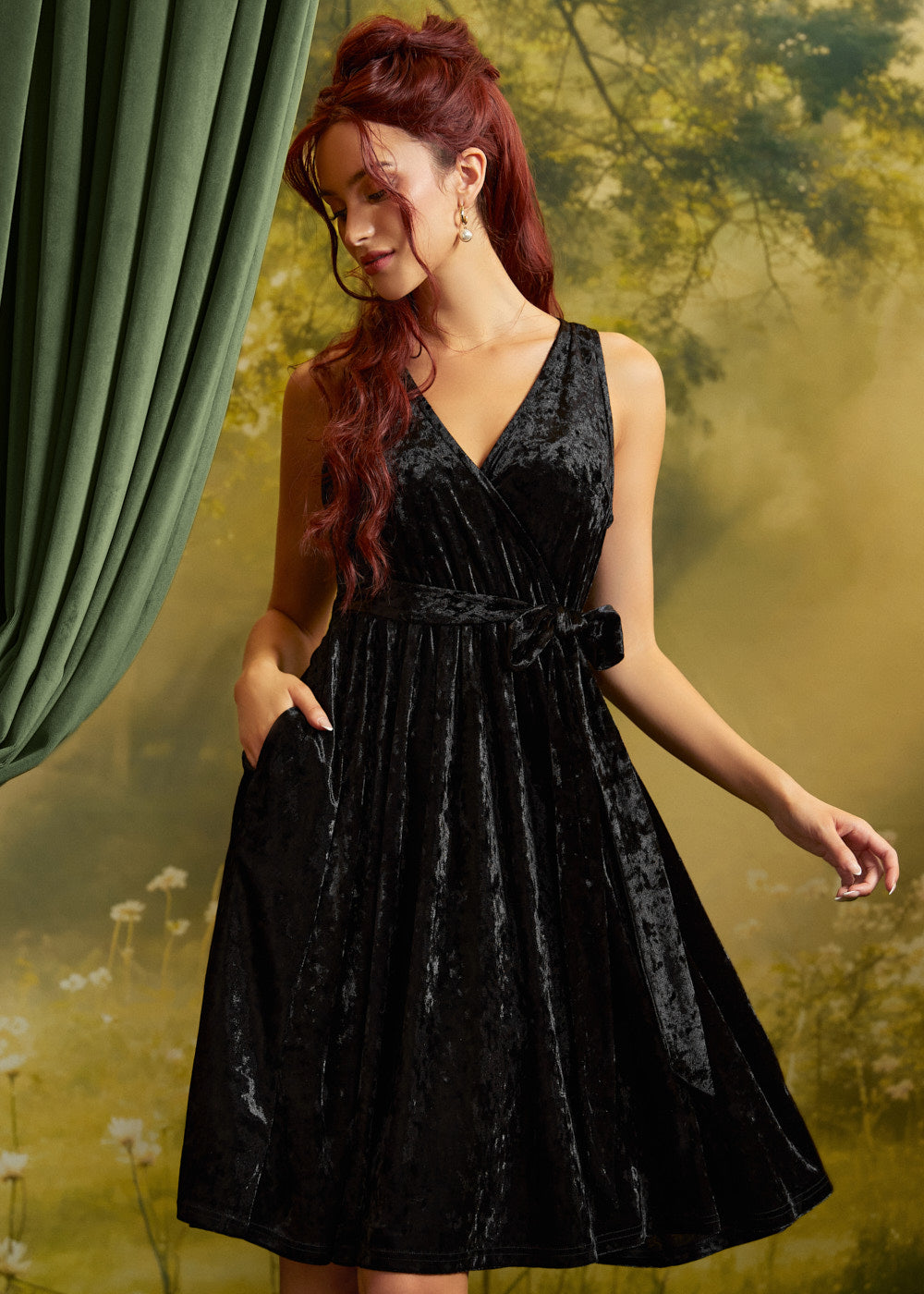 A woman with red hair wearing a sleeveless black velvet wrap dress with a tie waist and side pockets, posed against a soft-focus green and yellow backdrop with a green curtain on the left.
