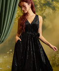 A woman with red hair wearing a sleeveless black velvet wrap dress with a tie waist and side pockets, posed against a soft-focus green and yellow backdrop with a green curtain on the left.