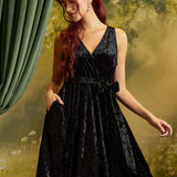 A woman with red hair wearing a sleeveless black velvet wrap dress with a tie waist and side pockets, posed against a soft-focus green and yellow backdrop with a green curtain on the left.
