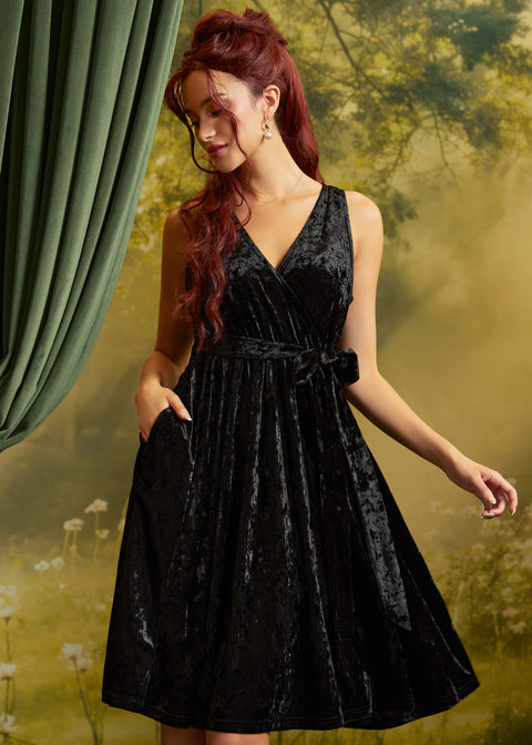 A woman with red hair wearing a sleeveless black velvet wrap dress with a tie waist and side pockets, posed against a soft-focus green and yellow backdrop with a green curtain on the left.
