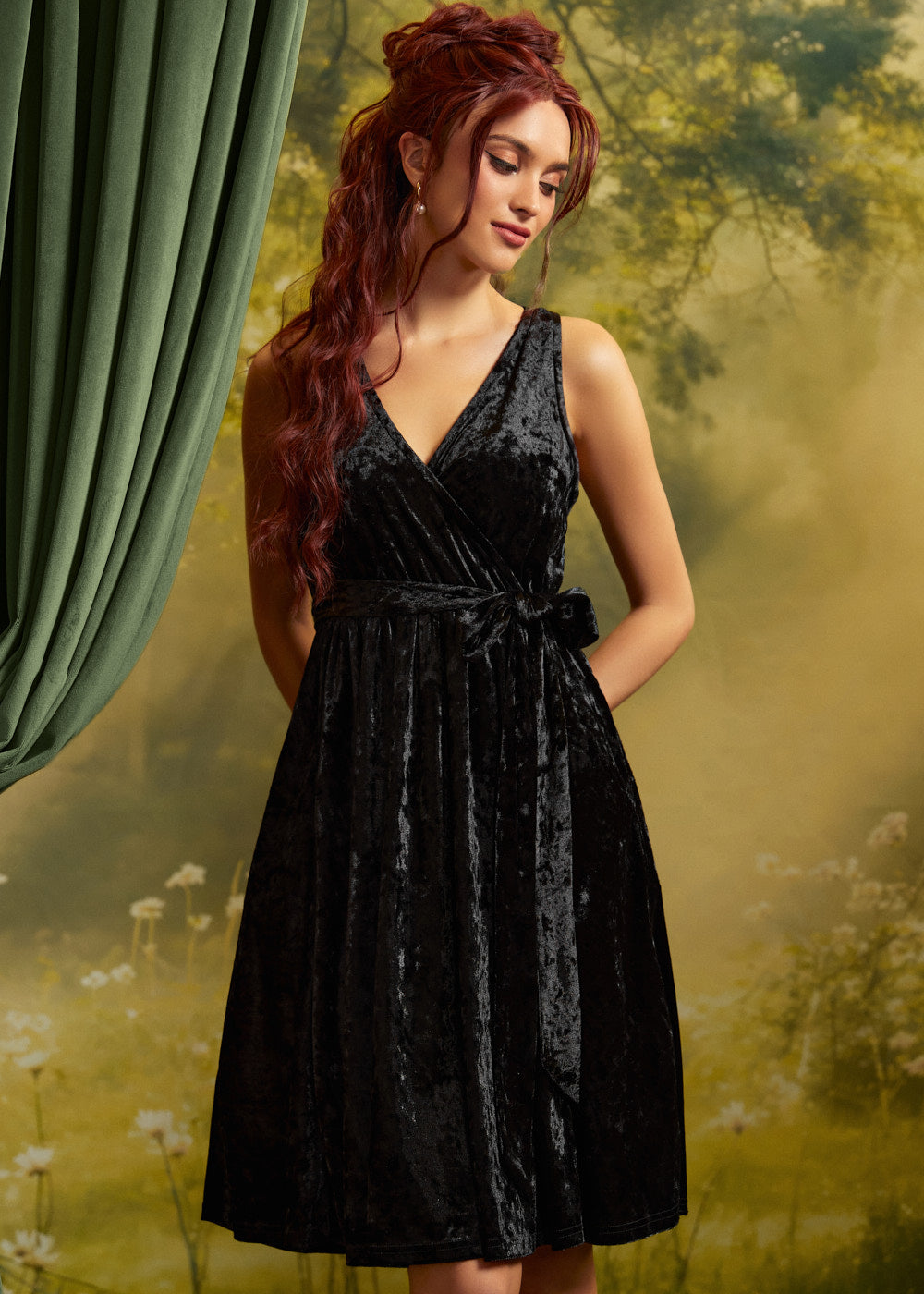Model wearing a sleeveless black crushed velvet faux-wrap dress with a V-neckline, matching tie waist, and softly gathered skirt against a garden-inspired backdrop.