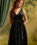 Model wearing a sleeveless black crushed velvet faux-wrap dress with a V-neckline, matching tie waist, and softly gathered skirt against a garden-inspired backdrop.