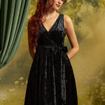 Model wearing a sleeveless black crushed velvet faux-wrap dress with a V-neckline, matching tie waist, and softly gathered skirt against a garden-inspired backdrop.