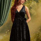 Model wearing a sleeveless black crushed velvet faux-wrap dress with a V-neckline, matching tie waist, and softly gathered skirt against a garden-inspired backdrop.