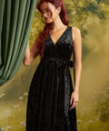A woman with long red hair models a sleeveless black velvet wrap dress by ModCloth with a V-neckline and tie waist, standing in front of a green curtain and a soft-focus yellow-green forest backdrop.