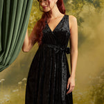 A woman with long red hair models a sleeveless black velvet wrap dress by ModCloth with a V-neckline and tie waist, standing in front of a green curtain and a soft-focus yellow-green forest backdrop.