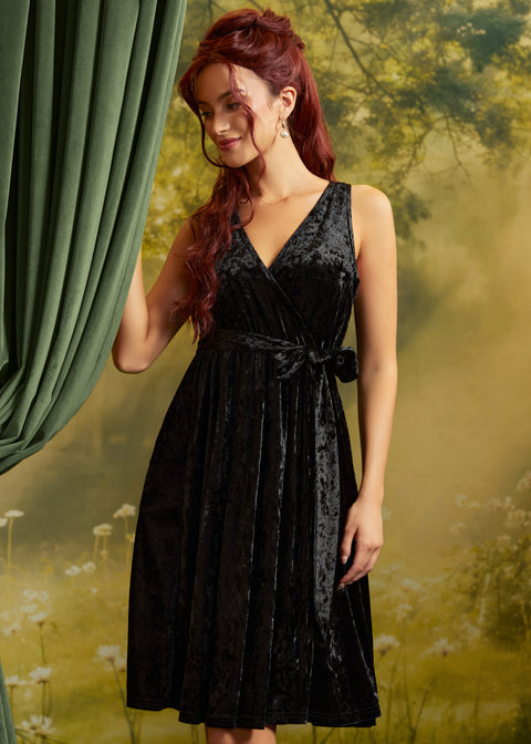 A woman with long red hair models a sleeveless black velvet wrap dress by ModCloth with a V-neckline and tie waist, standing in front of a green curtain and a soft-focus yellow-green forest backdrop.