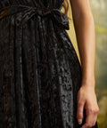 Close-up of a black crushed velvet dress showing its textured sheen, removable self-tie waist sash tied in a bow, and gathered skirt.