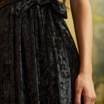 Close-up of a black crushed velvet dress showing its textured sheen, removable self-tie waist sash tied in a bow, and gathered skirt.