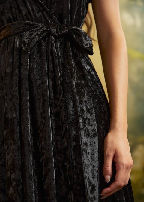 Close-up of a black crushed velvet dress showing its textured sheen, removable self-tie waist sash tied in a bow, and gathered skirt.
