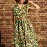 A woman wearing a green and brown patterned midi dress with a matching belt, posing in front of a rustic brick wall.