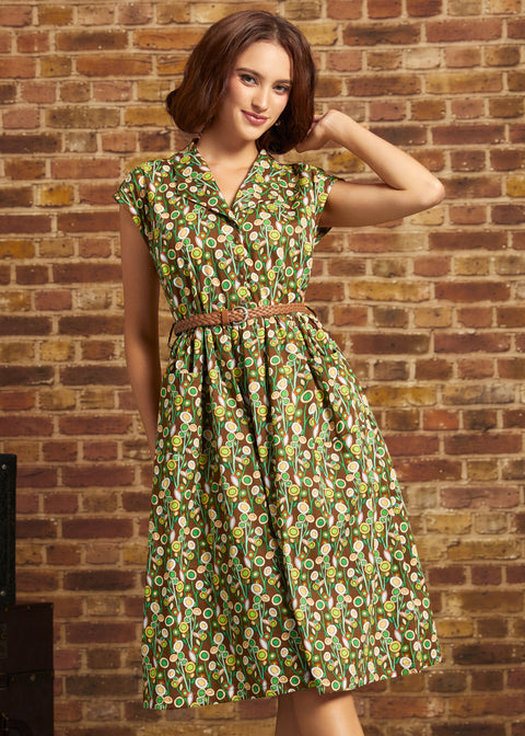 A woman wearing a green and brown patterned midi dress with a matching belt, posing in front of a rustic brick wall.
