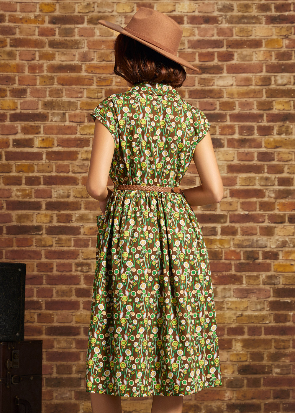 A woman viewed from behind wearing a green patterned midi dress with a brown belt and a brown wide-brim hat, standing against a brick wall.