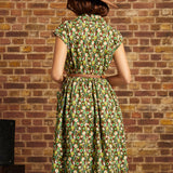 A woman viewed from behind wearing a green patterned midi dress with a brown belt and a brown wide-brim hat, standing against a brick wall.
