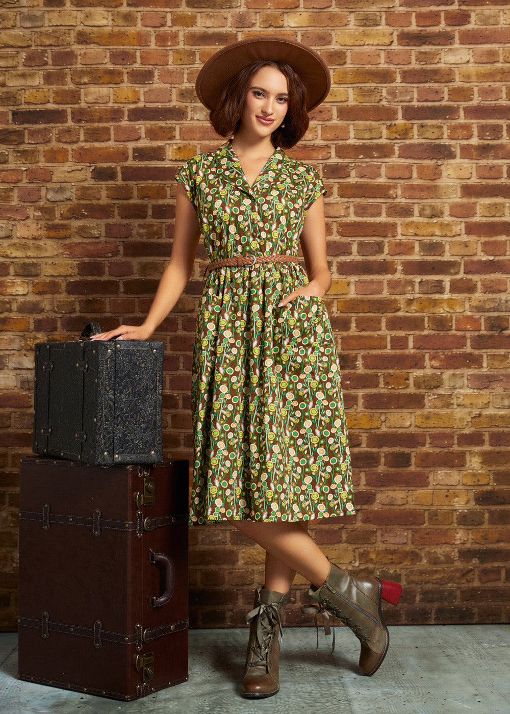 A woman stands in front of a brick wall wearing a green floral midi dress with a brown belt, a brown wide-brim hat, and brown lace-up boots. She is posed next to two stacked vintage-style trunks.