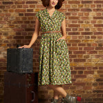 A woman stands in front of a brick wall wearing a green floral midi dress with a brown belt, a brown wide-brim hat, and brown lace-up boots. She is posed next to two stacked vintage-style trunks.