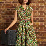 A woman wearing a green and yellow floral print midi dress with a brown belt and a wide-brimmed brown hat, standing next to a vintage black trunk against a brick wall.