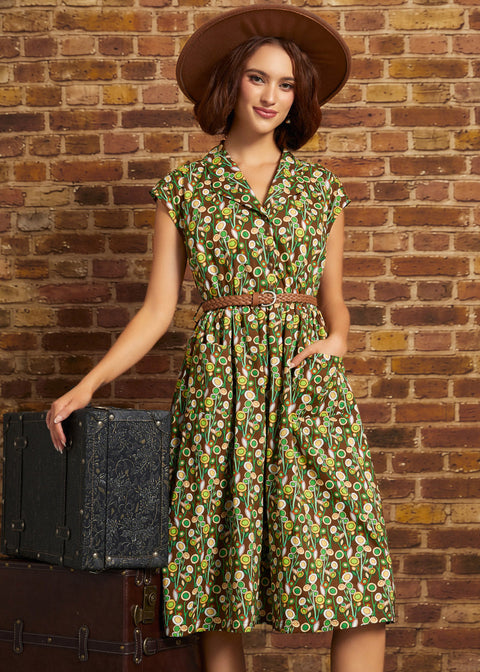A woman wearing a green and yellow floral print midi dress with a brown belt and a wide-brimmed brown hat, standing next to a vintage black trunk against a brick wall.