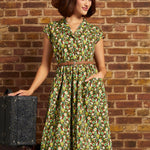 Model wearing the Card Catalog Cutie Dress in Olive Always, featuring a brown, green, and yellow retro floral print, braided belt, and flared skirt, styled with a brown hat.