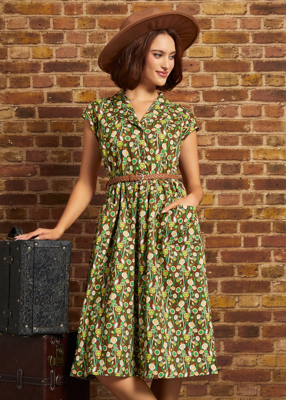 Model wearing the Card Catalog Cutie Dress in Olive Always, featuring a brown, green, and yellow retro floral print, braided belt, and flared skirt, styled with a brown hat.