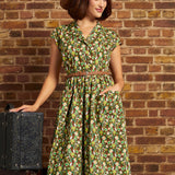 Model wearing the Card Catalog Cutie Dress in Olive Always, featuring a brown, green, and yellow retro floral print, braided belt, and flared skirt, styled with a brown hat.