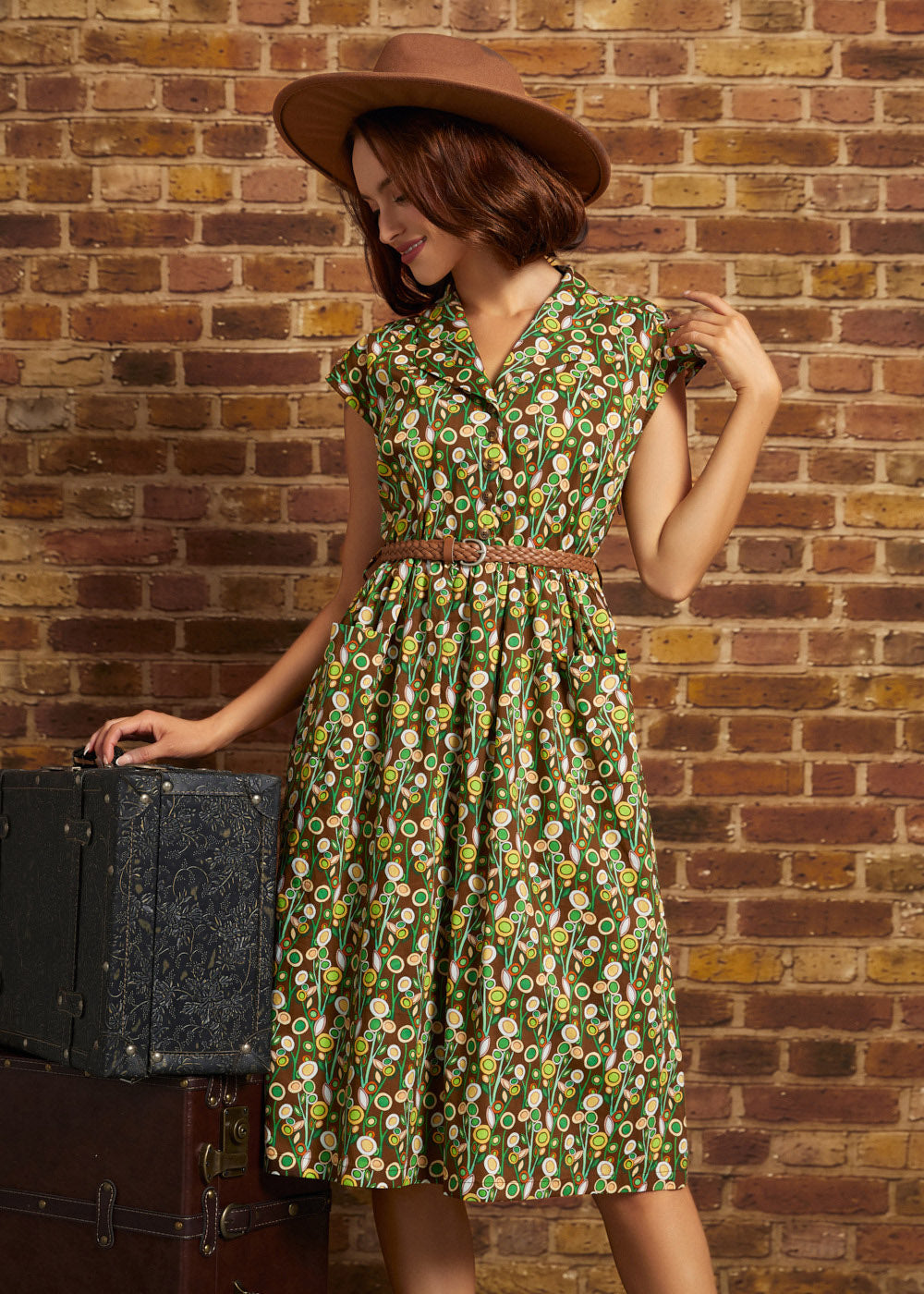 A woman wearing a brown wide-brim hat and a green floral print dress with a brown belt, posing next to a vintage black trunk against a brick wall.