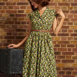 A woman wearing a brown wide-brim hat and a green floral print dress with a brown belt, posing next to a vintage black trunk against a brick wall.