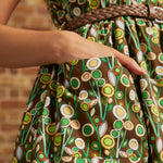 Close-up of a person wearing a brown dress with a retro floral pattern in green, yellow, and white, featuring a braided brown belt at the waist.
