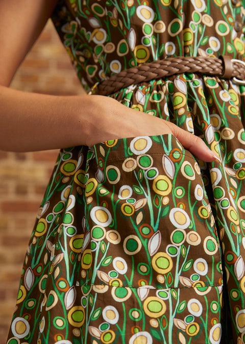 Close-up of a person wearing a brown dress with a retro floral pattern in green, yellow, and white, featuring a braided brown belt at the waist.