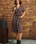 A woman stands in front of a brick wall wearing a short-sleeve plaid wrap dress with a tie waist, a gray beret, and dark lace-up ankle boots. A vintage trunk is visible on the left side of the scene.