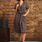 A woman stands in front of a brick wall wearing a short-sleeve plaid wrap dress with a tie waist, a gray beret, and dark lace-up ankle boots. A vintage trunk is visible on the left side of the scene.