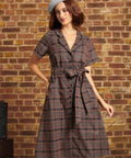 Model wearing the Going Out and About Dress in brown and black plaid with turquoise stripes, a button-front closure, matching tie belt, and gray beret against a brick wall.