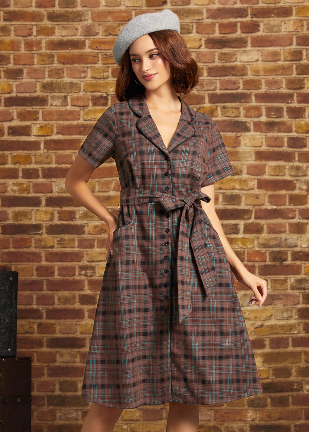Model wearing the Going Out and About Dress in brown and black plaid with turquoise stripes, a button-front closure, matching tie belt, and gray beret against a brick wall.