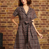 Model wearing the Going Out and About Dress in brown and black plaid with turquoise stripes, a button-front closure, matching tie belt, and gray beret against a brick wall.