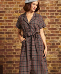 A woman wearing a short-sleeve plaid belted dress and a gray beret stands against a brick wall background.