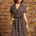 A woman wearing a short-sleeve plaid belted dress and a gray beret stands against a brick wall background.