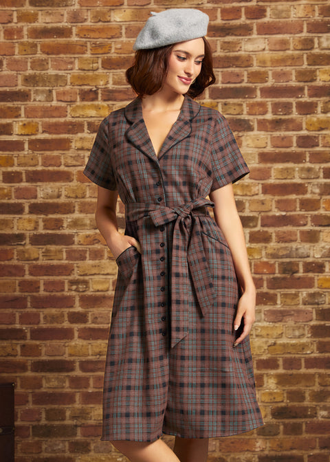 A woman wearing a short-sleeve plaid belted dress and a gray beret stands against a brick wall background.