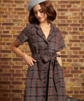 A woman wearing a short-sleeve plaid dress with a tie waist and a gray beret, standing against a brick wall.
