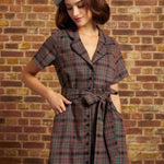 A woman wearing a short-sleeve plaid dress with a tie waist and a gray beret, standing against a brick wall.