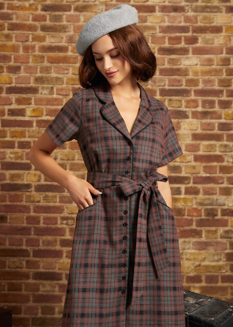 A woman wearing a short-sleeve plaid dress with a tie waist and a gray beret, standing against a brick wall.
