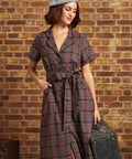 A woman stands against a brick wall wearing a short-sleeve plaid ModCloth dress with a tie waist, a gray beret, and holding a vintage suitcase.