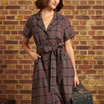 A woman stands against a brick wall wearing a short-sleeve plaid ModCloth dress with a tie waist, a gray beret, and holding a vintage suitcase.
