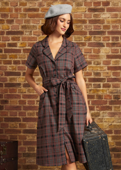 A woman stands against a brick wall wearing a short-sleeve plaid ModCloth dress with a tie waist, a gray beret, and holding a vintage suitcase.