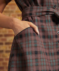 Close-up of a dress's front pocket with black contrast piping, showing a hand tucked inside and the brown plaid fabric with turquoise windowpane stripes.